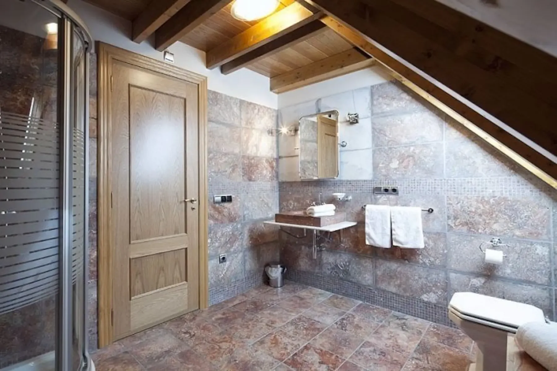 Attic bathroom with sloped wooden ceiling, tiled walls and floor, shower cabin, wooden door, wall-mounted sink, mirror, and toilet.