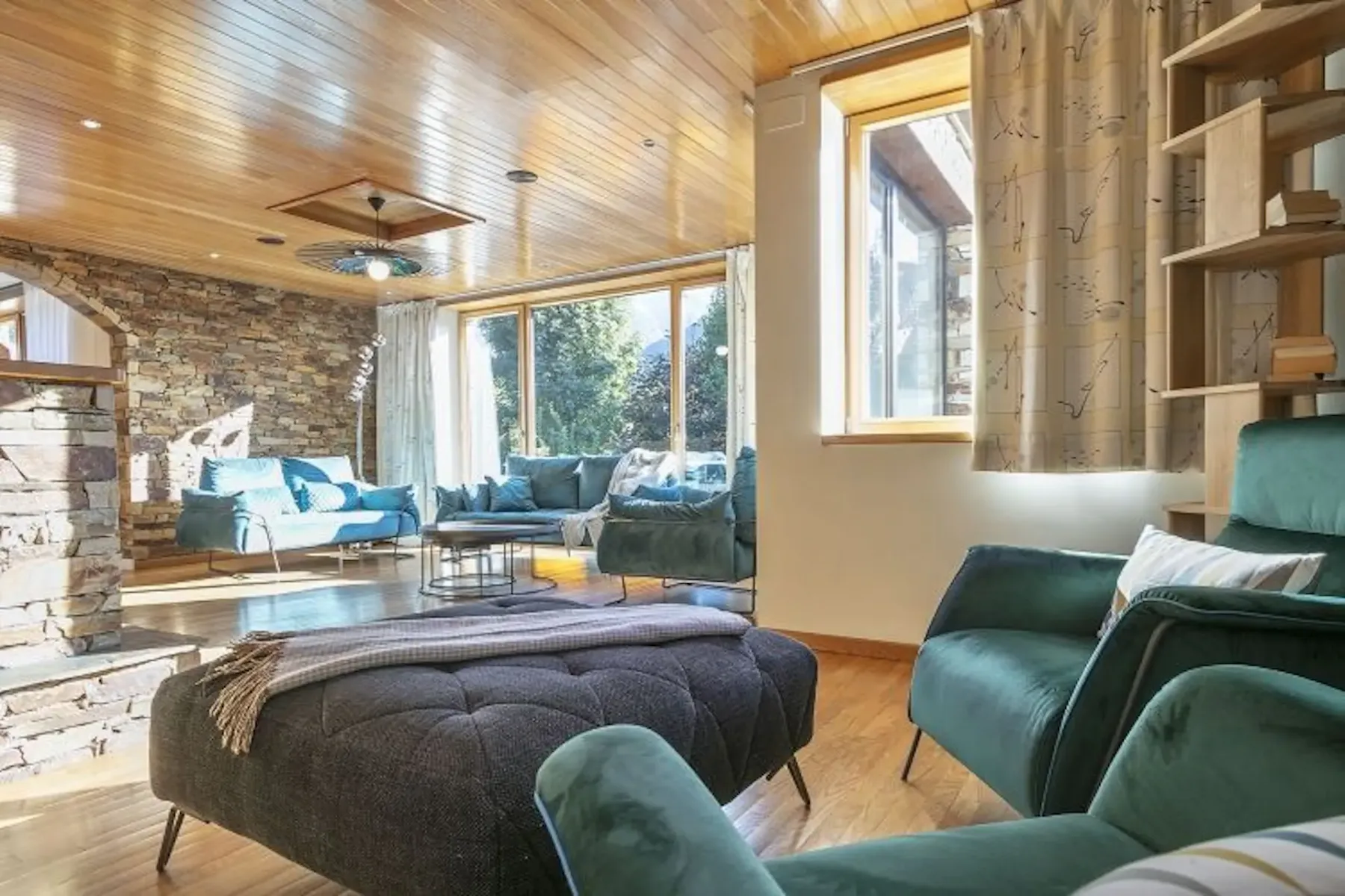 A sunlit living room with stone walls, wooden ceiling, large windows, and teal upholstered furniture. A dark ottoman is in the center.