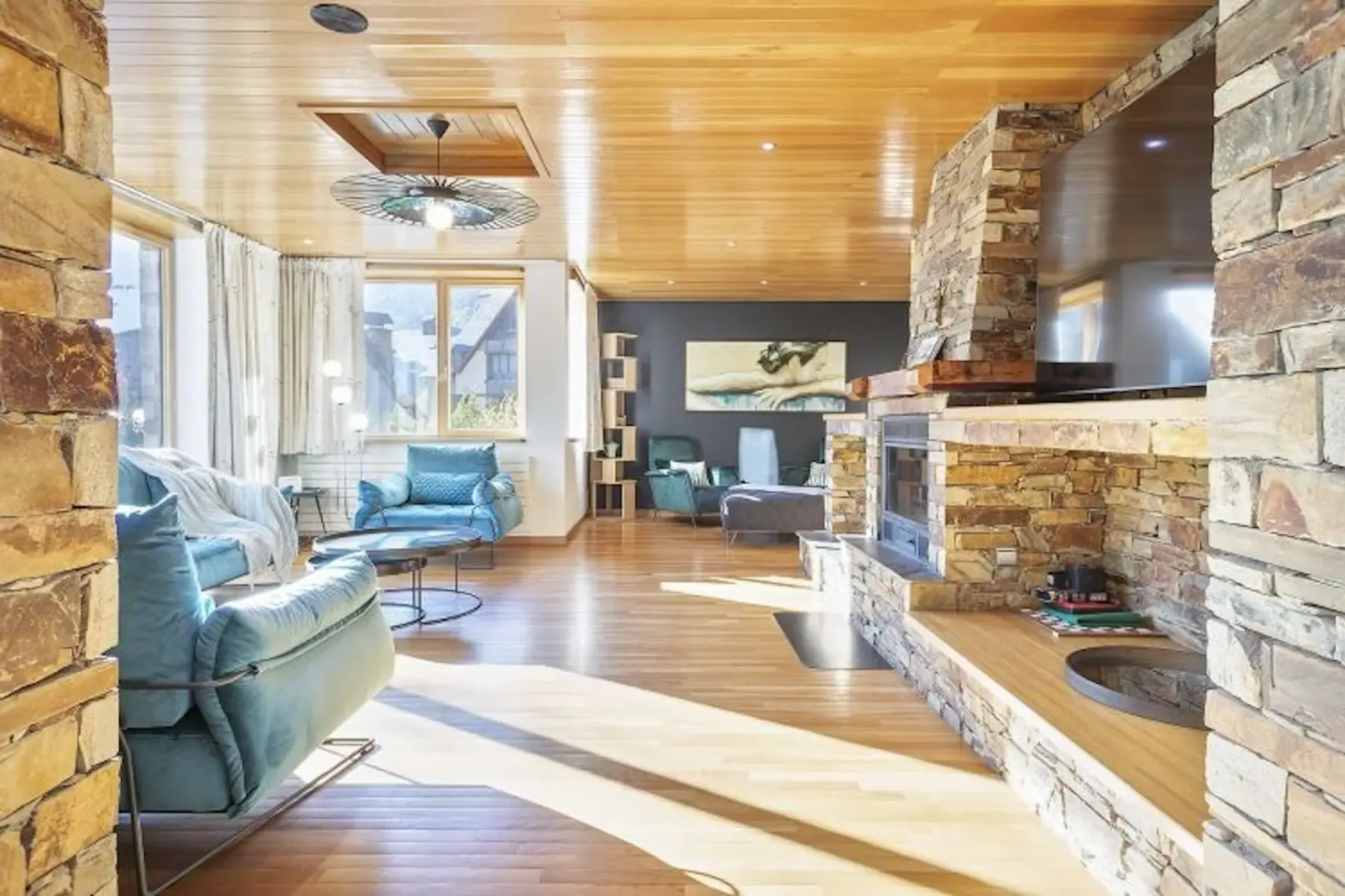 A spacious living room with stone walls, wooden ceiling, and large windows. It has blue armchairs, a central stone fireplace, and wooden floors, with artwork on the wall.
