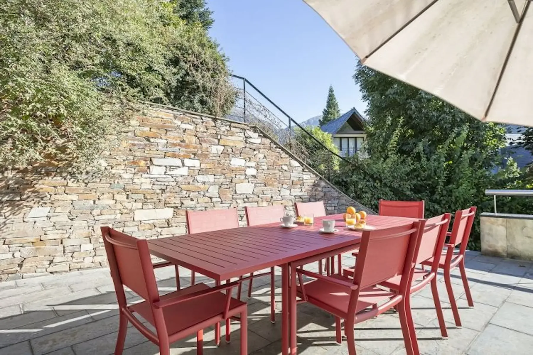 Red outdoor dining set with eight chairs on a stone patio, under a large umbrella. Stone wall and lush greenery in the background. Cups and a small pitcher are on the table.