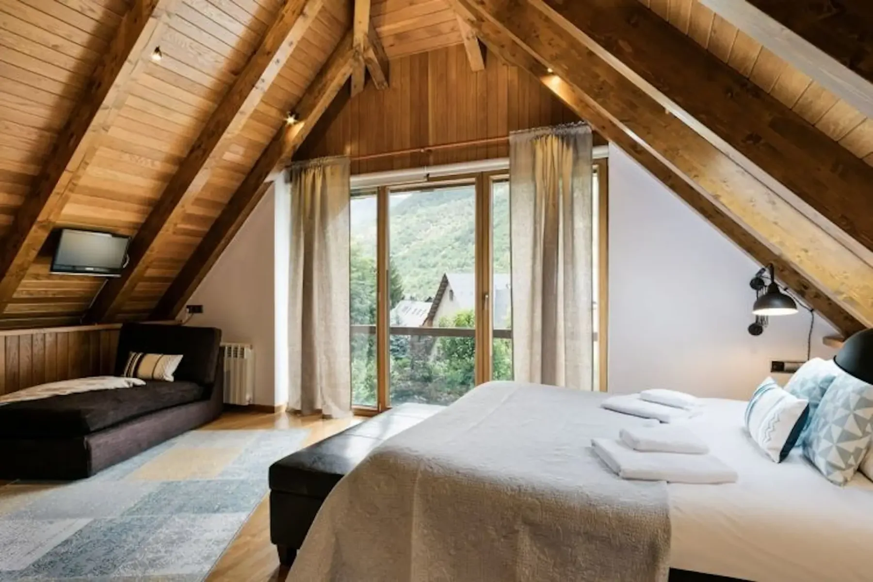 An attic bedroom with exposed wooden beams, two beds, and a glass door opening onto greenery.