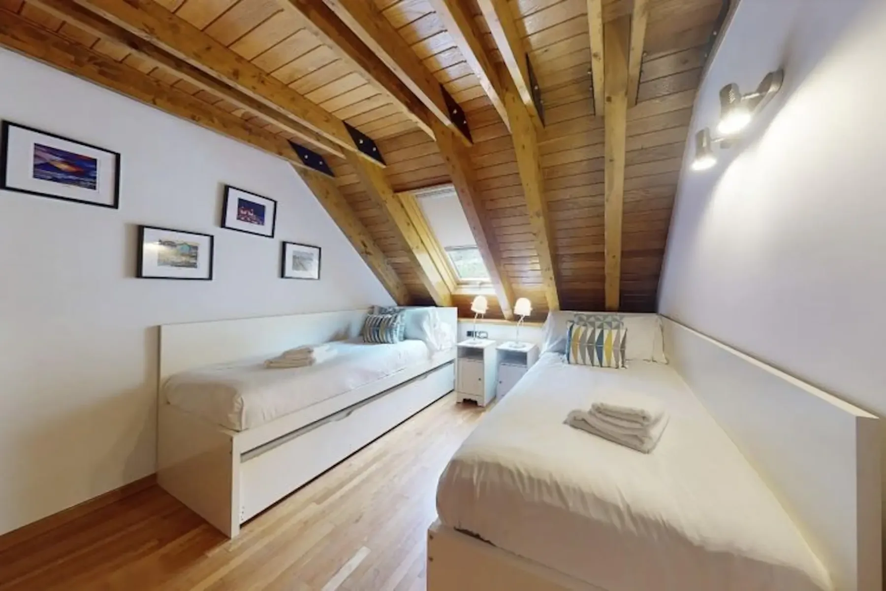 An attic bedroom with two single beds, a skylight, framed pictures on the walls, and a wooden ceiling. A small nightstand is between the beds, each topped with folded towels.