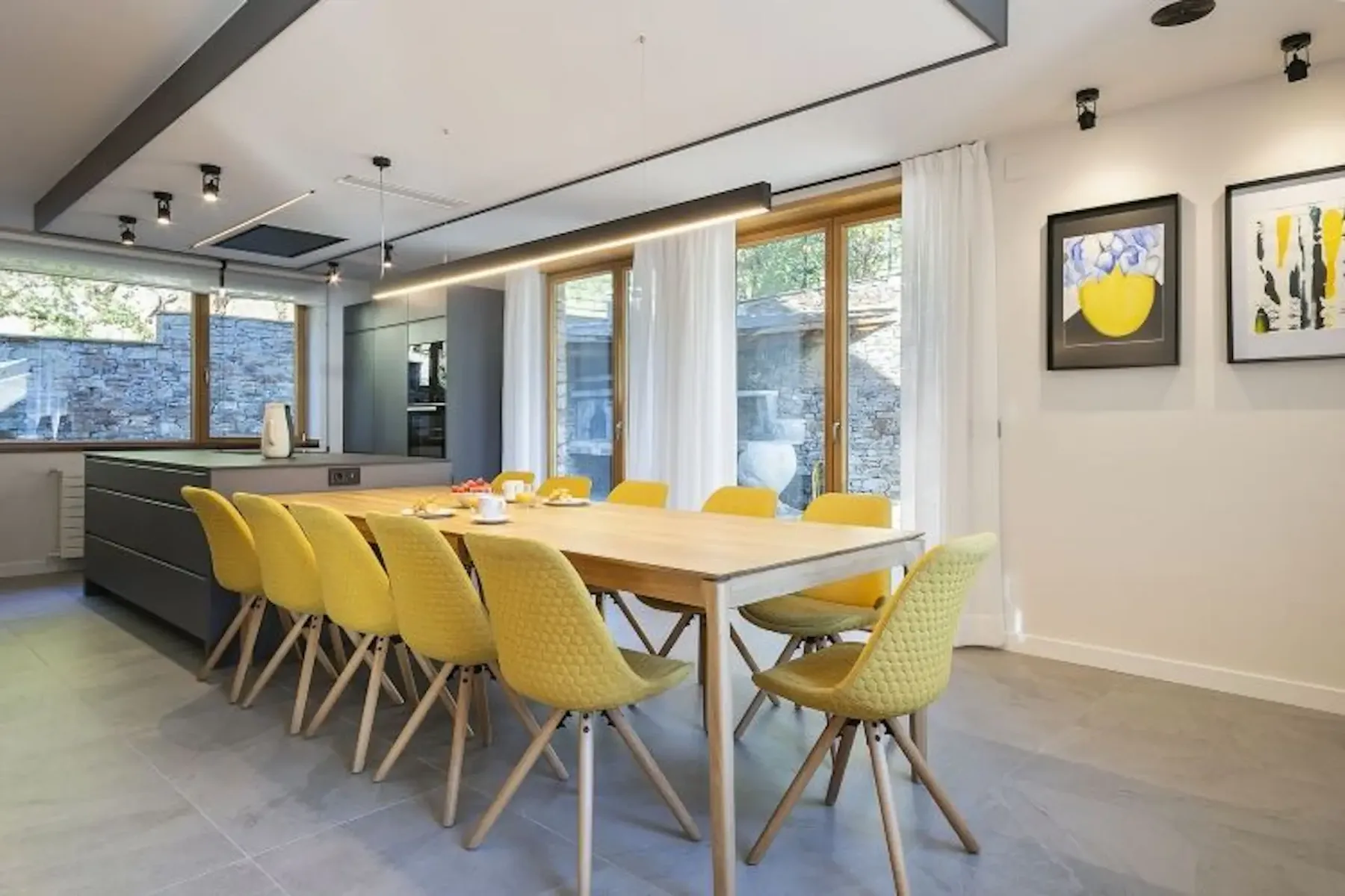 Dining room with a wooden table and eight yellow chairs, set for breakfast. Modern decor features artwork, large windows, gray cabinetry, and a view of a brick wall outside.