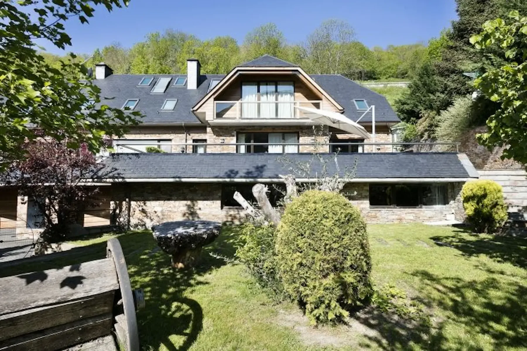 A large stone and wood house with multiple floors and a balcony is surrounded by a grassy yard with trees and shrubs.