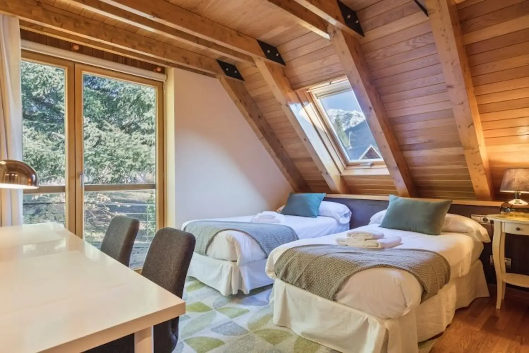 A cozy attic bedroom with two single beds, a skylight, wooden walls, a desk with chairs, and a window showing trees and mountains outside.