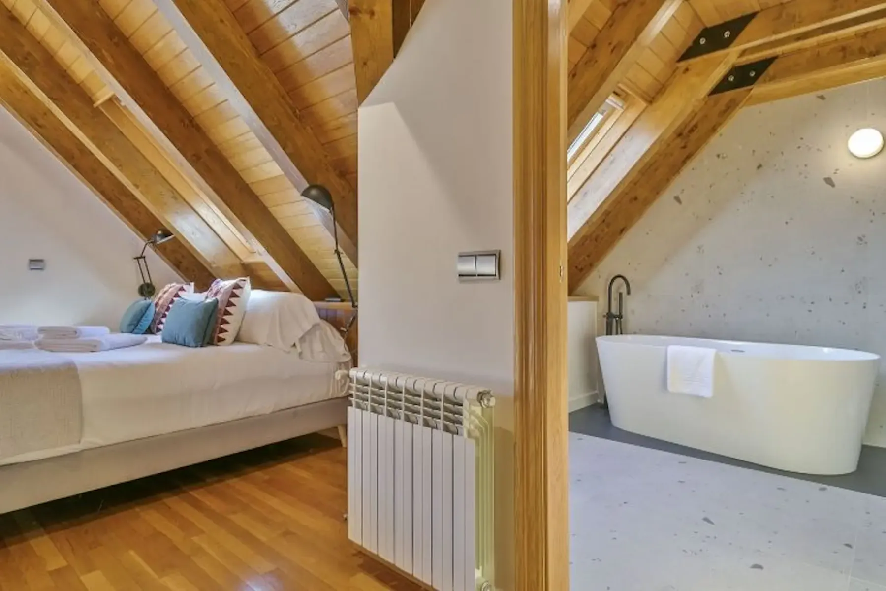 Attic room with exposed wooden beams, featuring a bed with pillows and a freestanding bathtub in an adjacent bathroom.