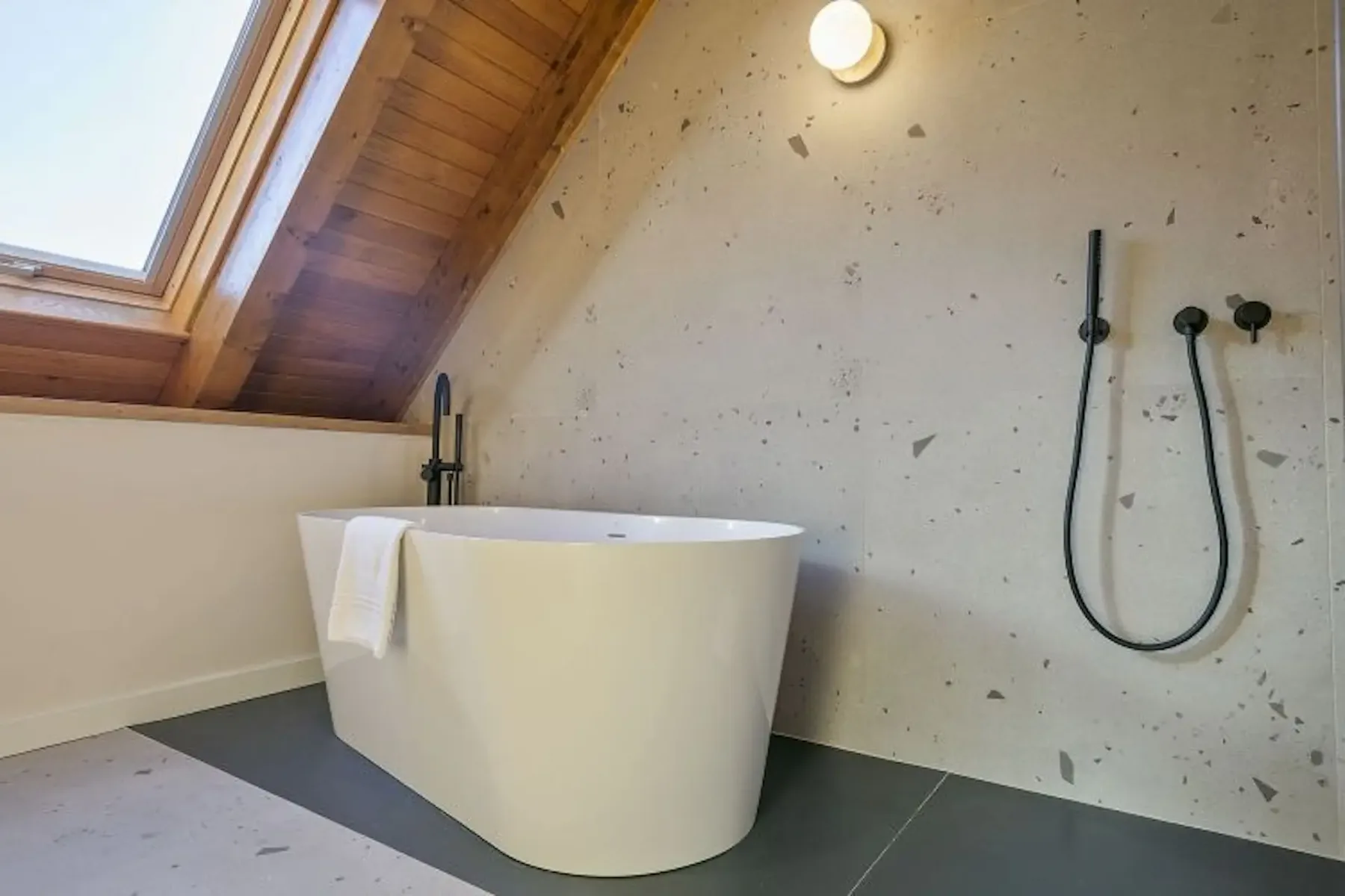 A modern bathroom features a freestanding white bathtub with a towel, sloped wooden ceiling, skylight, and wall-mounted black shower fixtures against a speckled wall.