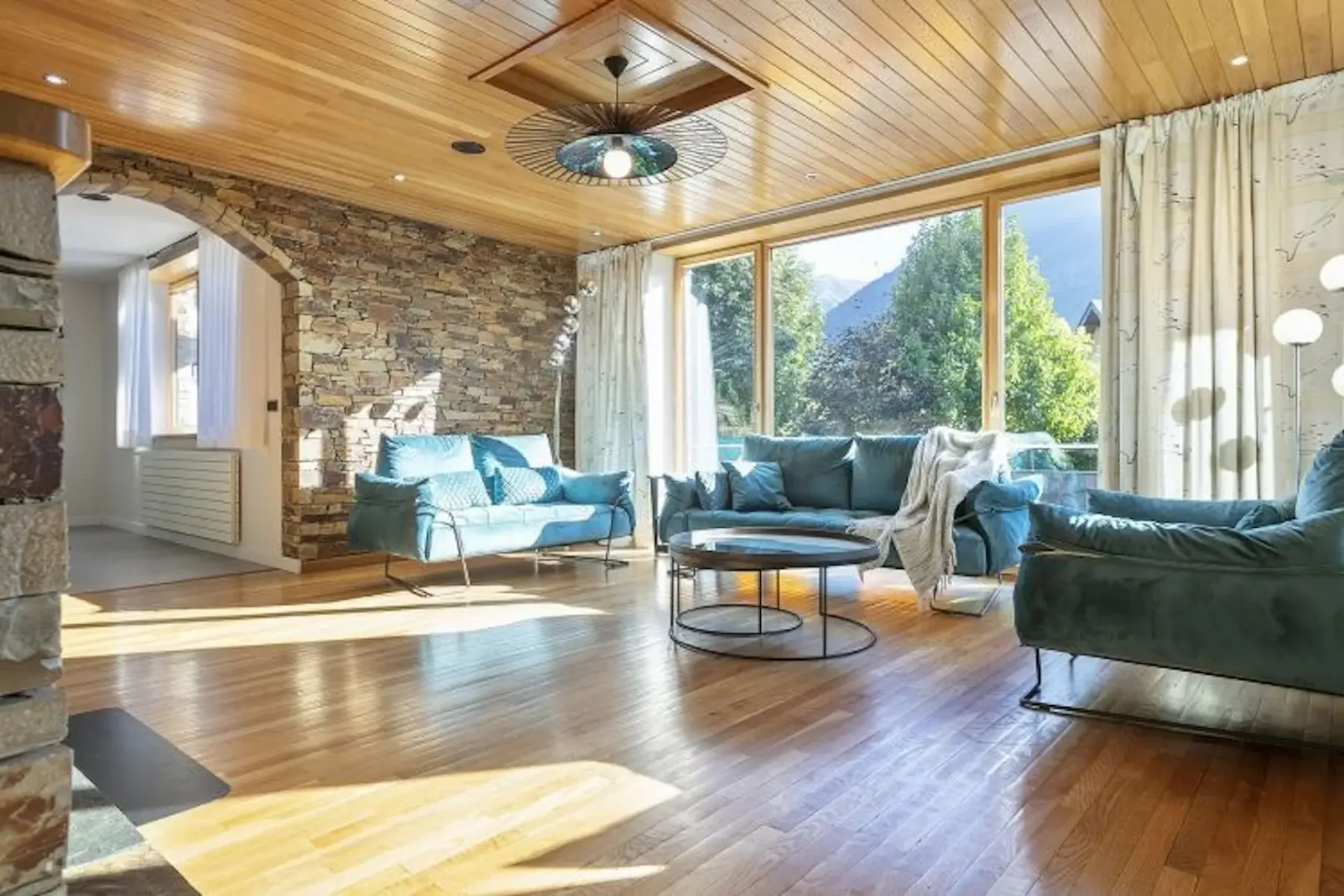 A spacious living room with stone walls, wooden ceiling, large windows, teal sofas, and a round coffee table. Sunlight streams in, creating a warm atmosphere.