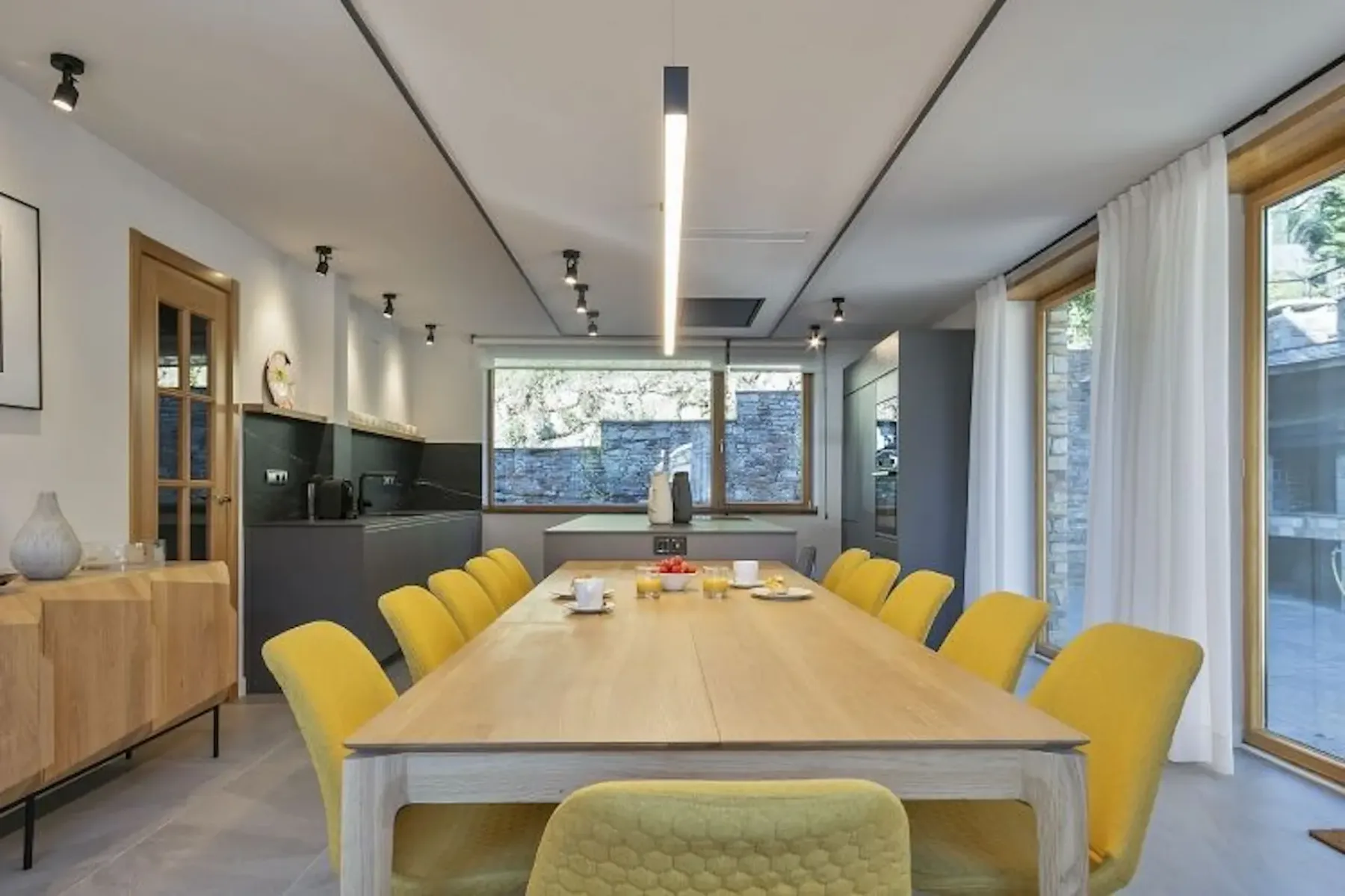 Modern kitchen and dining area with a wooden table, eight yellow chairs, and gray cabinets. A large window provides natural light, and a wooden sideboard is on the left.