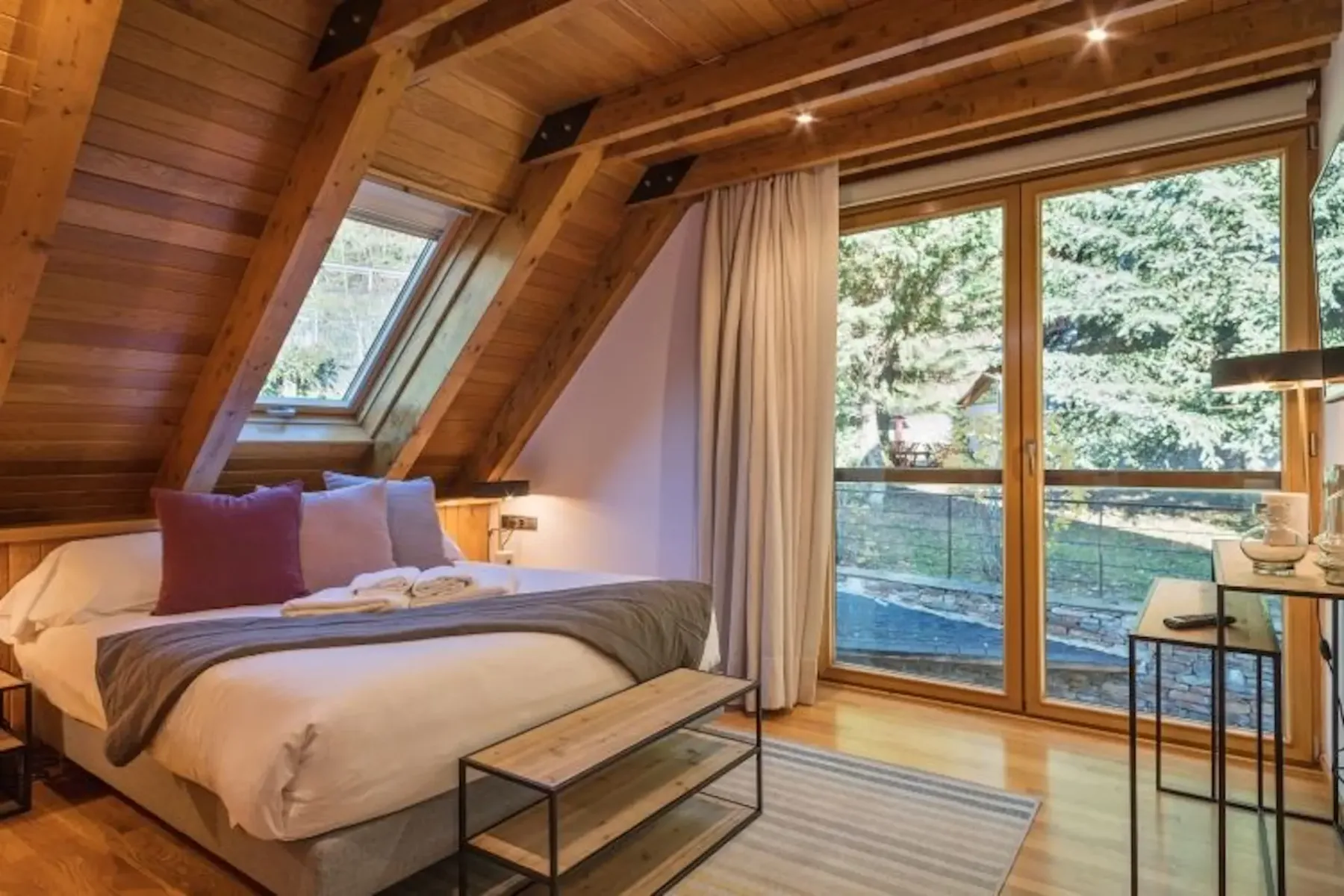 Cozy attic bedroom with wood paneling, a skylight, a large window with a view of trees, a double bed with pillows and blankets, a small table, and a striped rug on the floor.