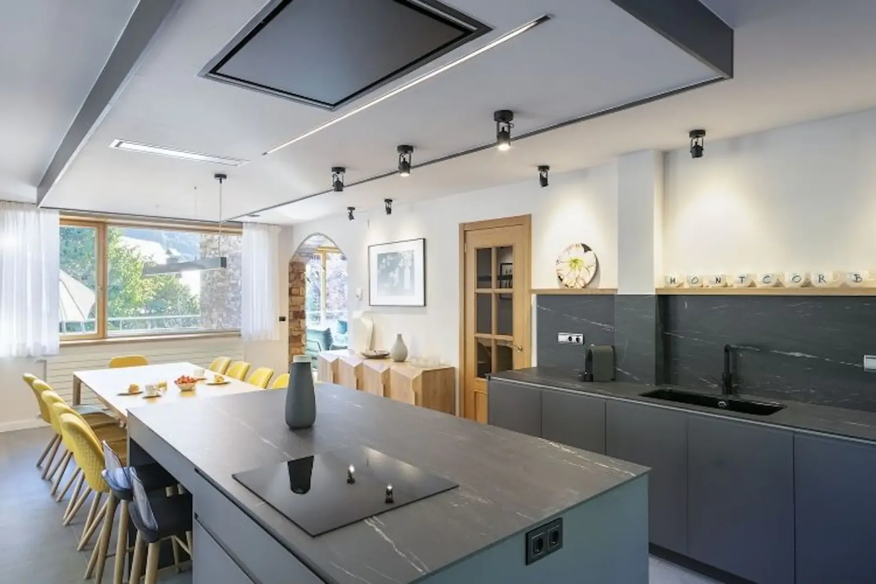 Modern kitchen with a gray island countertop, yellow chairs around a dining table, black backsplash, wooden door, wall decorations, and a large window with sheer curtains.