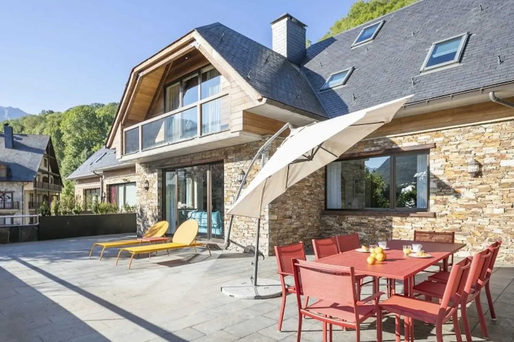 A modern house with stone facade, large windows, and a spacious patio. The patio features a red dining set, two yellow loungers, and a large umbrella under a clear blue sky.