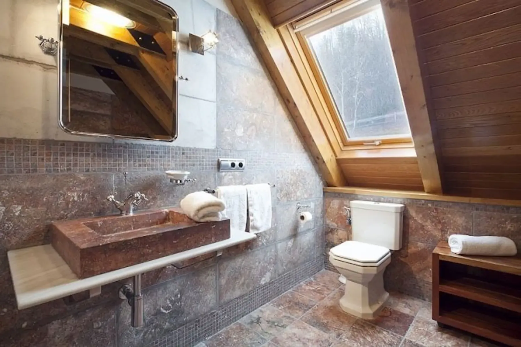 Bathroom with stone sink, wall-mounted mirror, toilet, and sloped wooden ceiling. Skylight window provides natural light. Towel rack and wooden shelf are nearby.