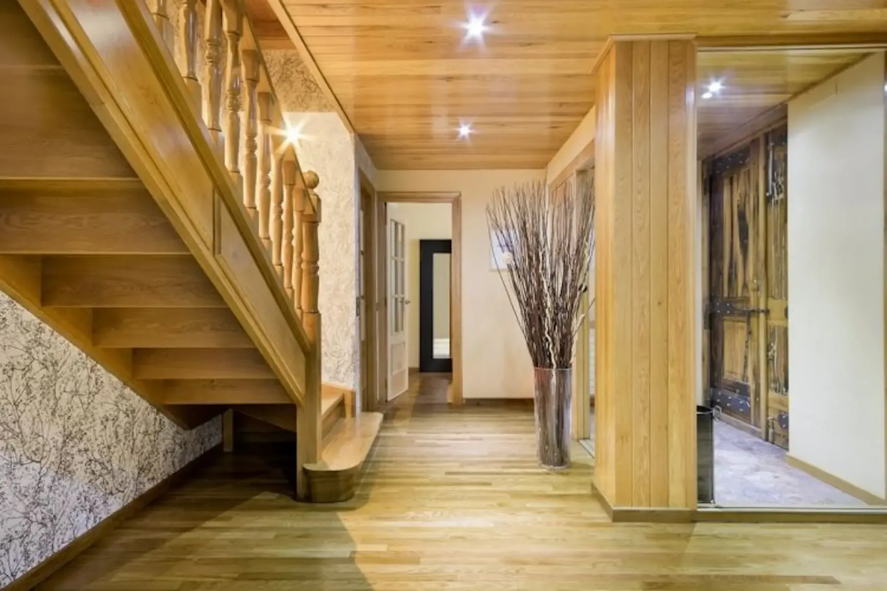 A wooden staircase rises beside a hallway with a tall vase of branches.