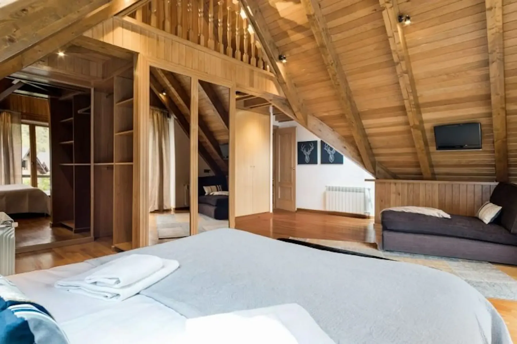 Cozy attic bedroom with wooden ceiling, featuring a bed, sofa, wall-mounted TV, and wardrobe with mirrored doors.