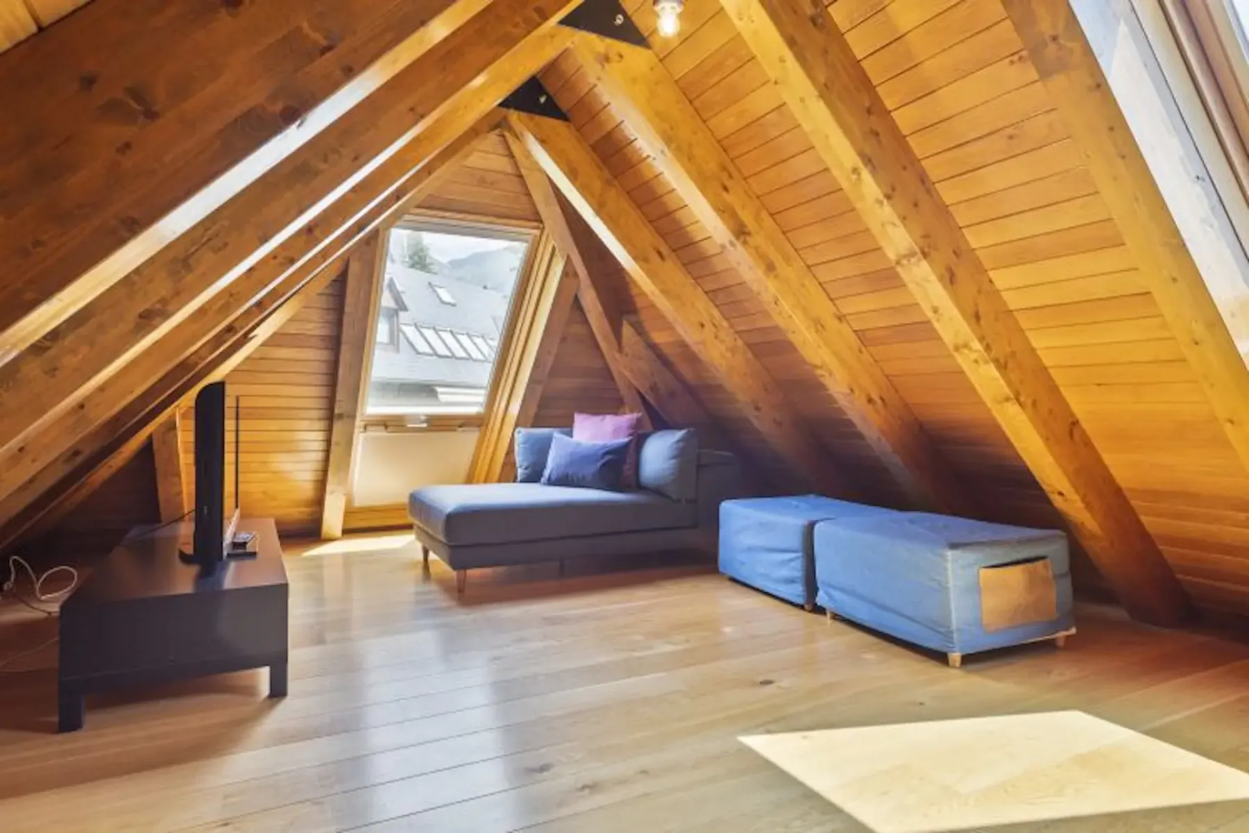A-frame attic with wooden beams and flooring, featuring a skylight, a small TV on a stand, a couch with pillows, and a blue ottoman.