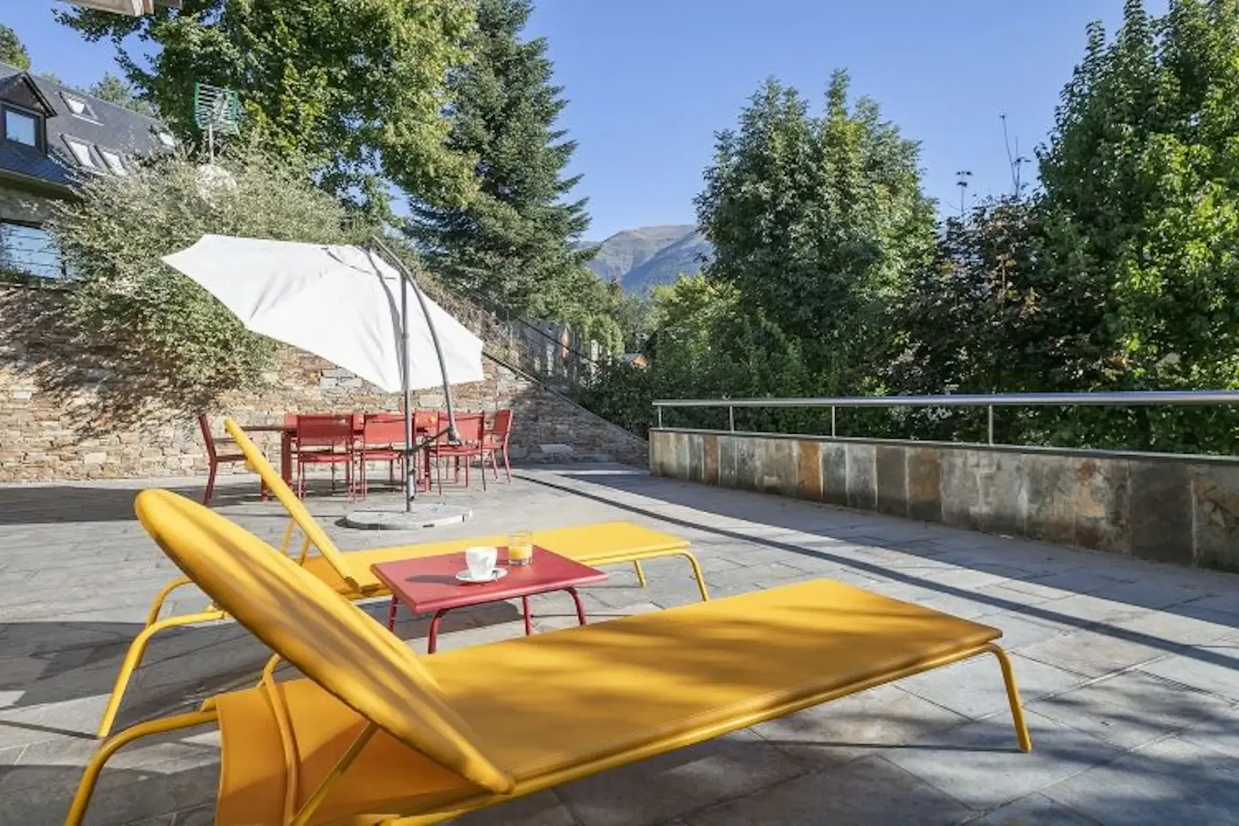 Outdoor patio with yellow lounge chairs, a red table with a cup, and a dining set under a white umbrella. Surrounded by trees and stone walls under a clear blue sky.