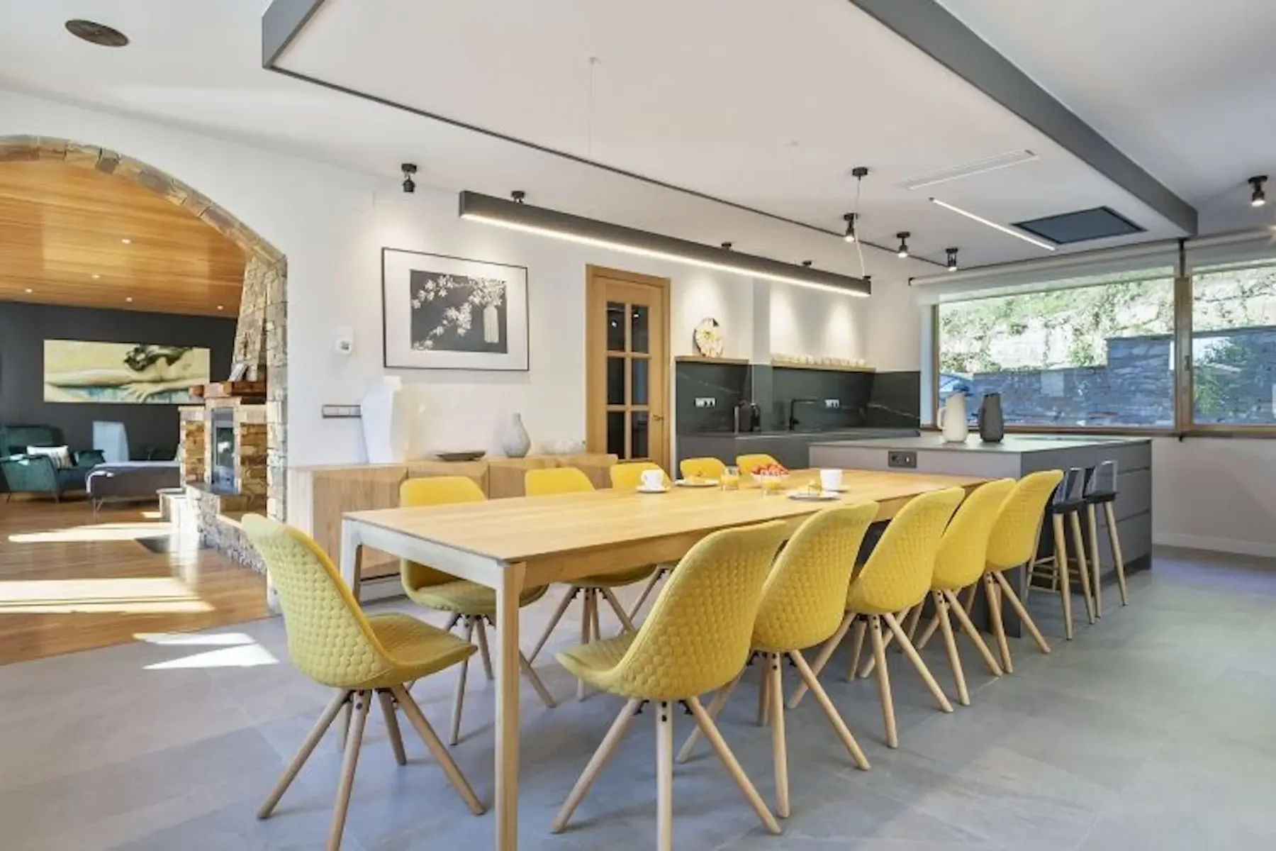 Modern kitchen and dining area with a long wooden table, yellow chairs, and sleek cabinets. A living room with a sofa and art on the wall is visible through a stone archway.