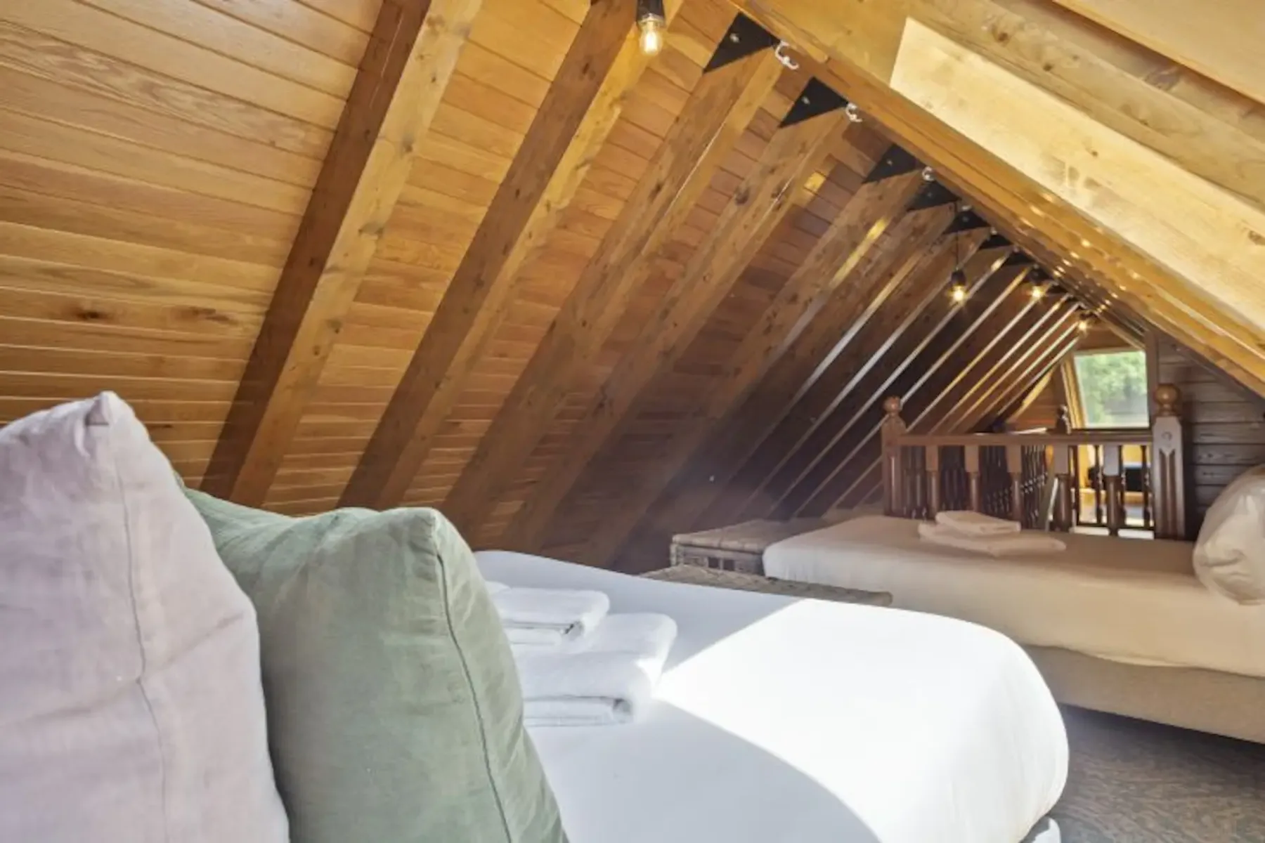 Cozy loft area with wooden beams, two beds with pillows and towels, and a small seating area. Natural light filters in from a window, creating a warm, inviting atmosphere.
