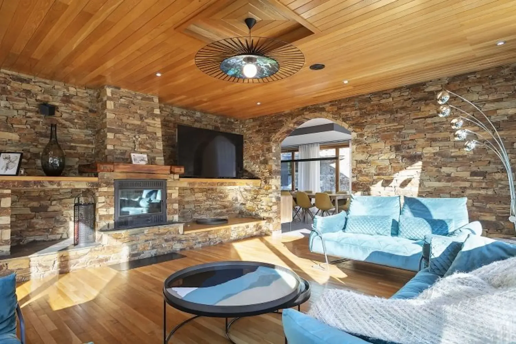 Living room with stone walls, wooden ceiling, and floor. Features a fireplace, wall-mounted TV, blue sofas, and modern lighting. An archway leads to a dining area.