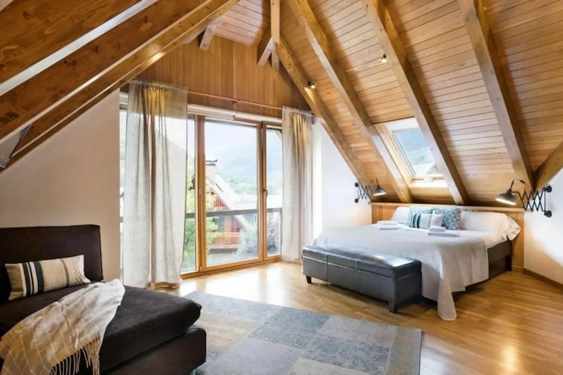 A cozy attic bedroom with a wooden ceiling, large windows, a double bed, and a chaise lounge. Natural light streams in, highlighting a rug on the wooden floor.