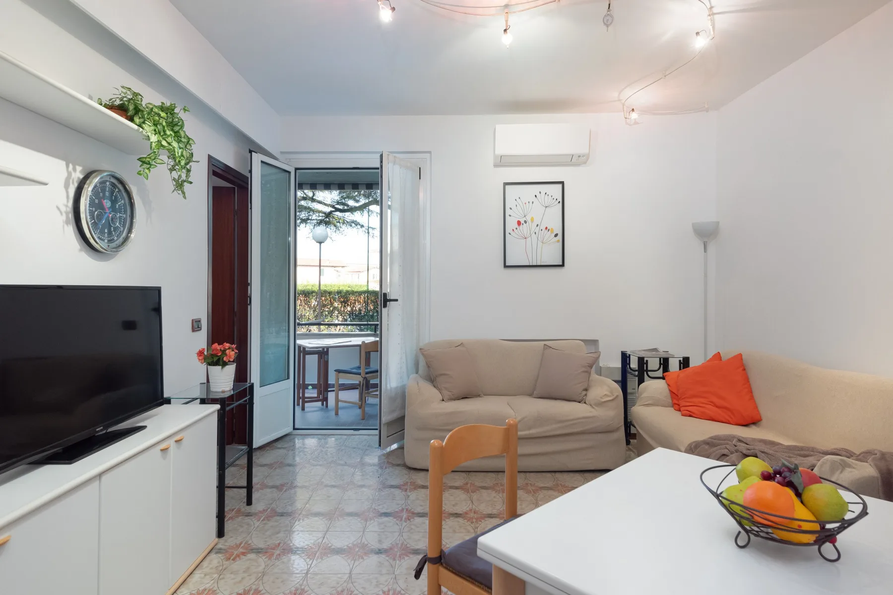 Una sala de estar con asientos, un televisor y una puerta abierta hacia un balcón.