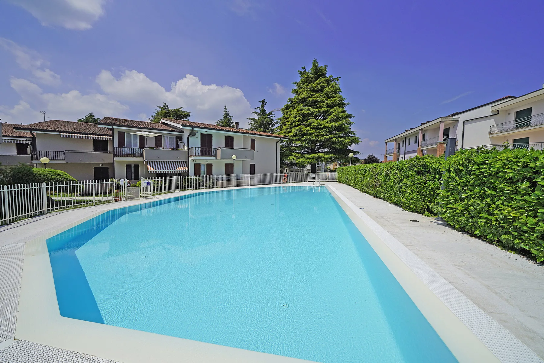 Una piscina al aire libre está junto a edificios residenciales y un seto.
