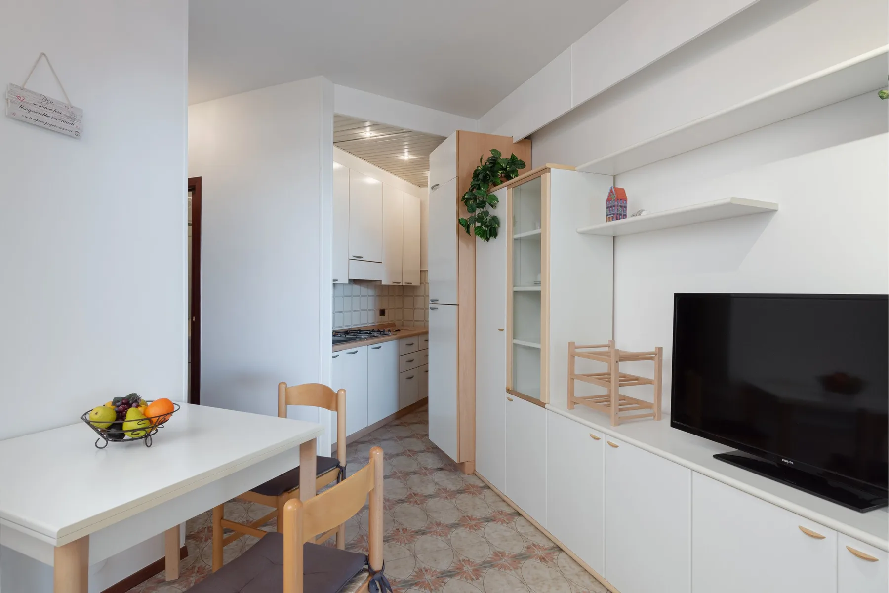 Una luminosa zona de comedor y estar con un televisor y una pequeña cocina al fondo.