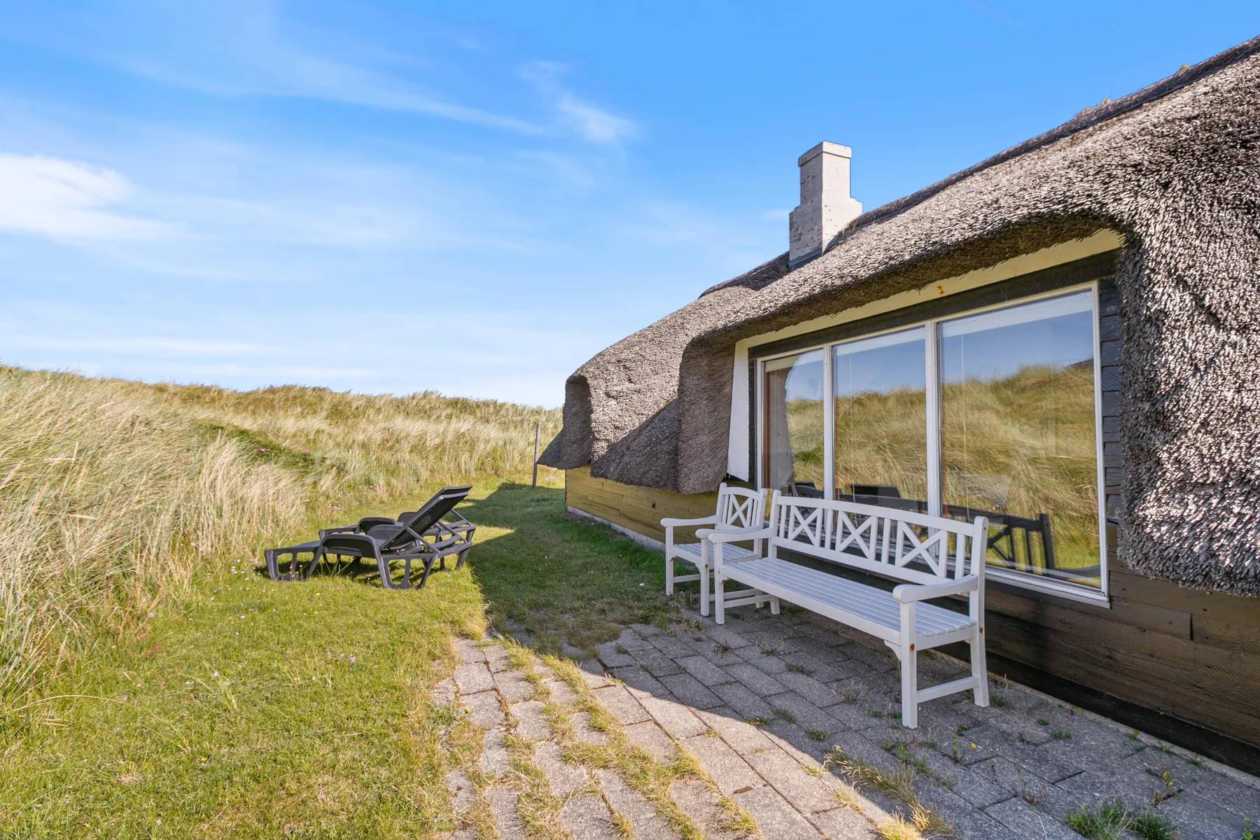 Una casa con techo de paja y asientos al aire libre junto a dunas y un patio pavimentado.