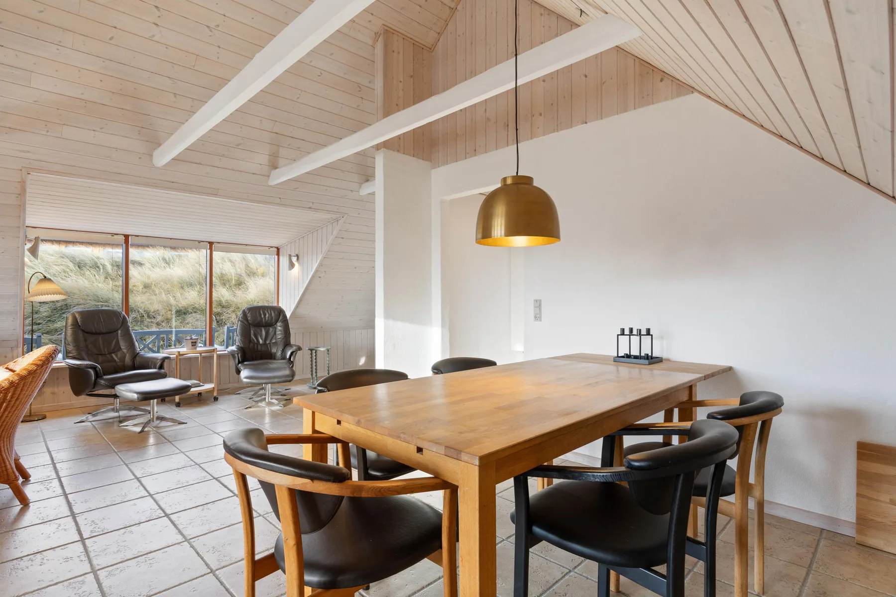 Comedor con mesa de madera y asientos, abierto a una sala de estar con grandes ventanas.