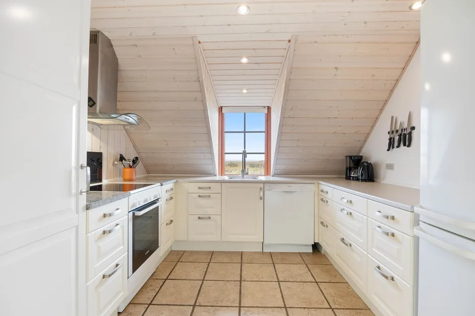 Una cocina luminosa con armarios blancos, suelo de baldosas y una ventana bajo el techo inclinado.