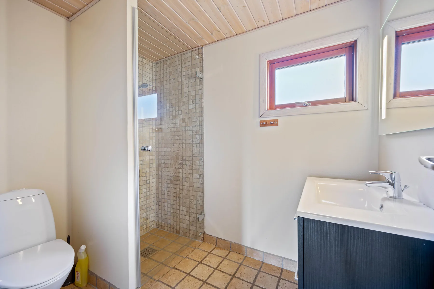 Un baño con ducha alicatada, inodoro y lavabo bajo ventanas pequeñas.