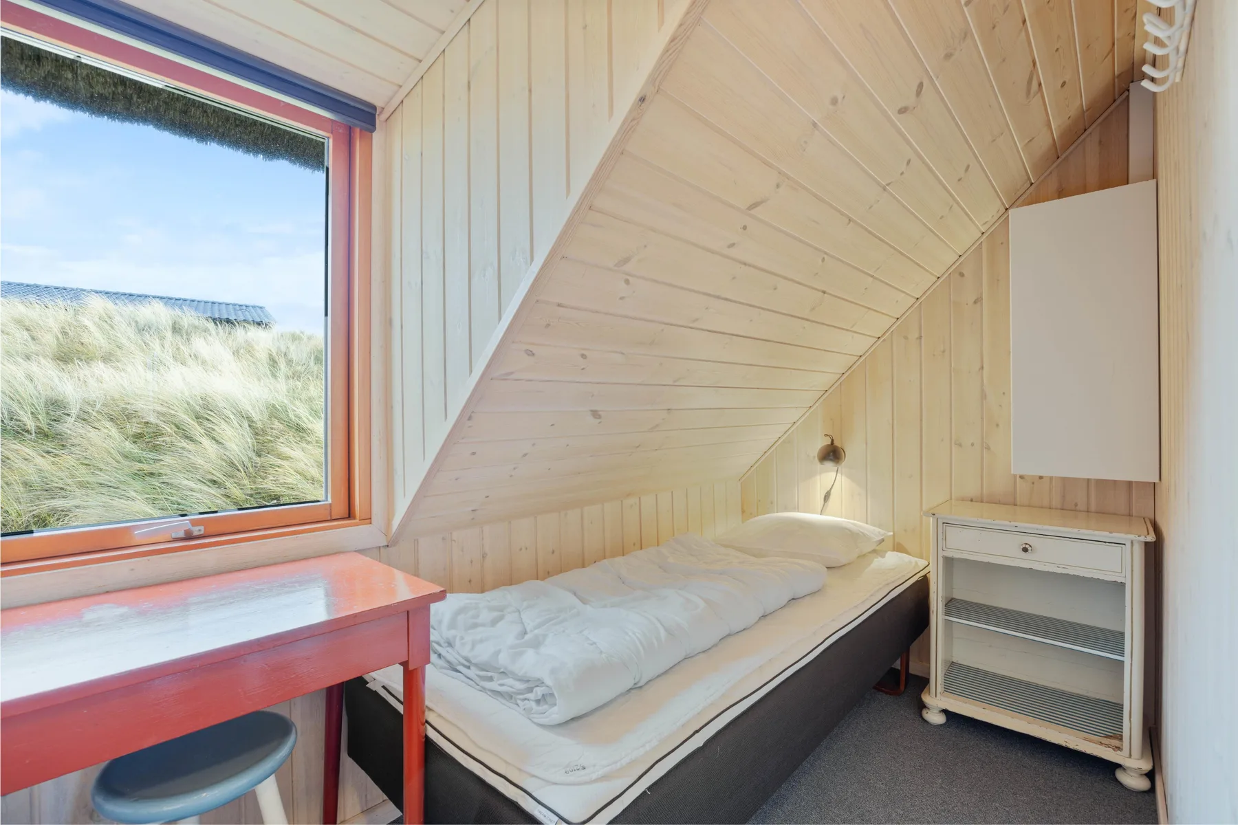 Un dormitorio con una cama bajo un techo inclinado y una ventana con vistas a dunas cubiertas de hierba.