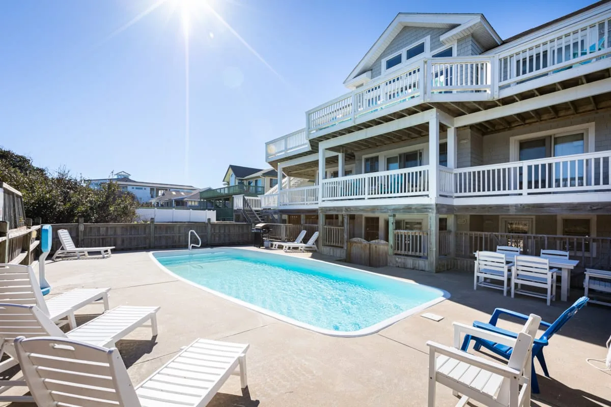 Una piscina ocupa un patio soleado junto a una casa de varios niveles con balcones blancos.