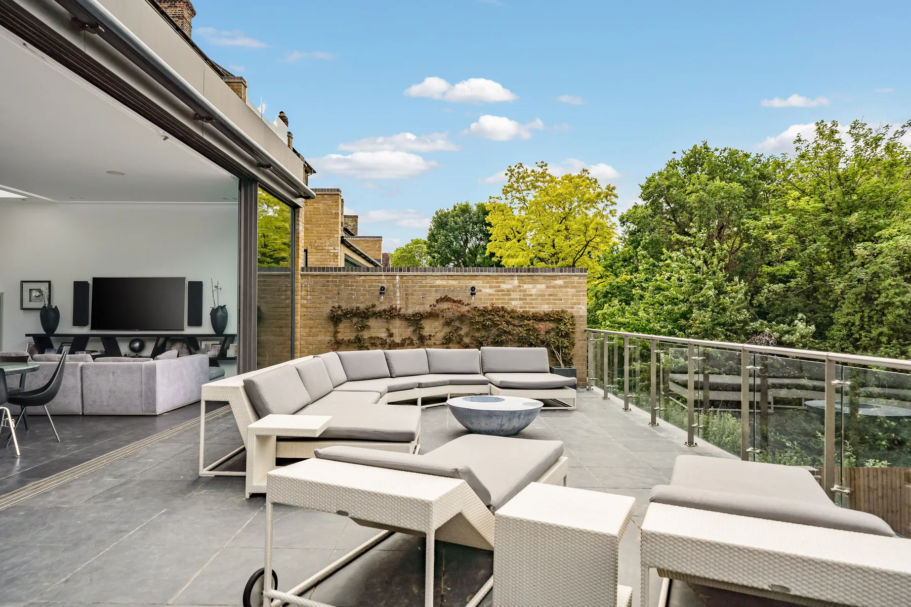 A rooftop terrace has outdoor seating beside glass railings and a leafy view.