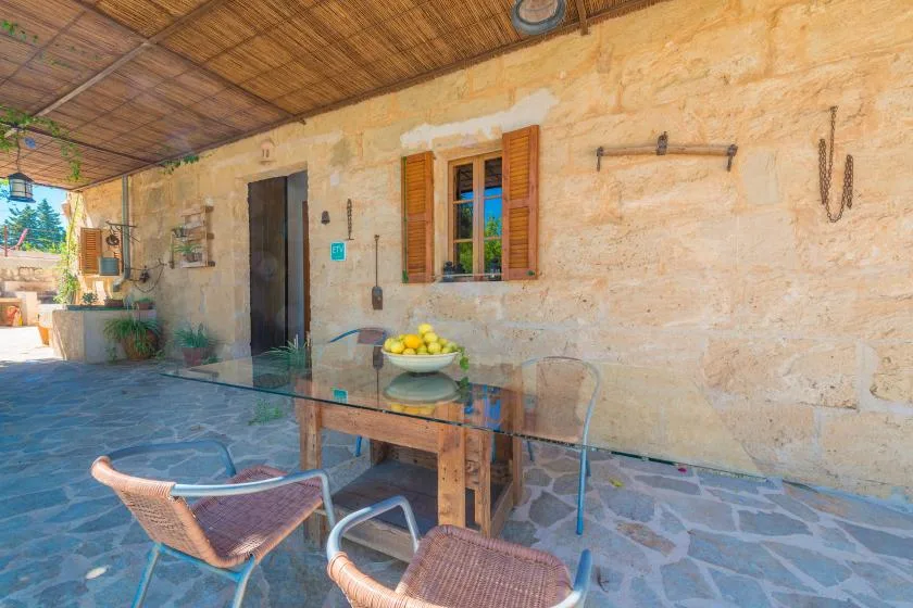A covered stone patio with a table, seating, and a shuttered window.