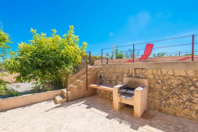 A sunny stone patio with an outdoor grill beside a leafy tree.