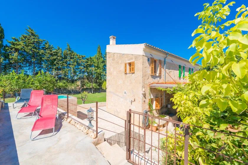 A stone house beside a sunny patio with red lounge seating and a pool.