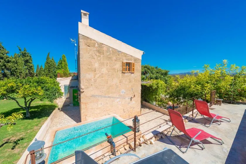 A small outdoor pool sits beside a stone building, with red lounge chairs on the patio.