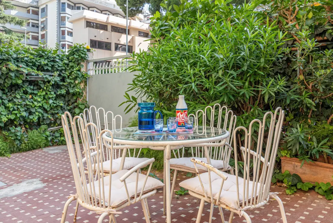 Un patio al aire libre con una mesa redonda y sillas rodeadas de plantas frondosas.