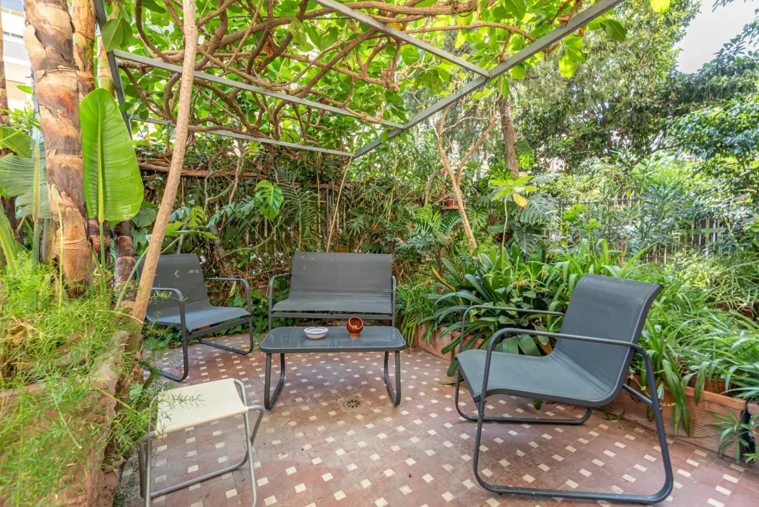 Un patio exterior frondoso tiene asientos alrededor de una mesa baja bajo una estructura cubierta de enredaderas.