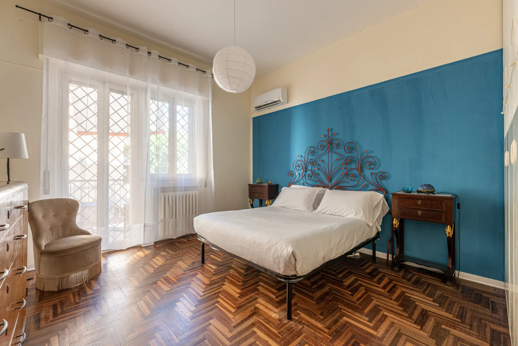 Un dormitorio con una cama blanca junto a una pared azul y ventanas altas.