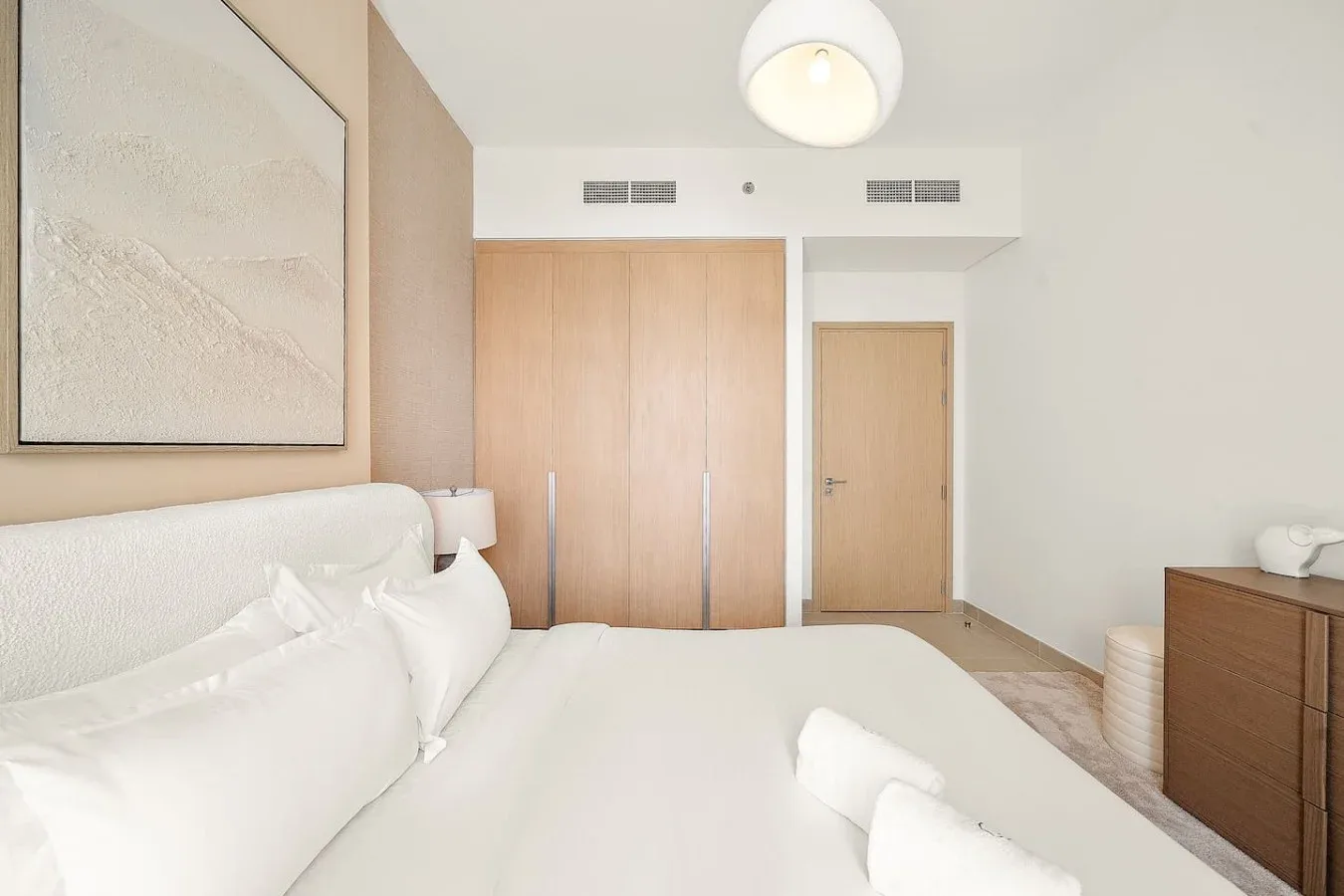A bright bedroom with a large bed, pale wood wardrobes, and a bedside wall light.
