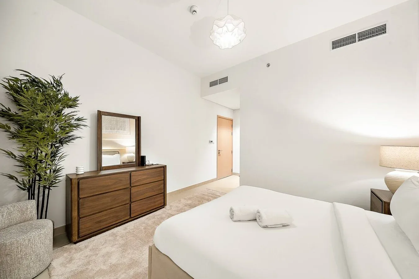 A bright bedroom with a large bed, wooden dresser, and leafy plant.
