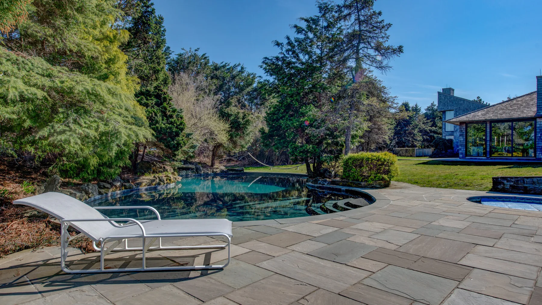 Una piscina al aire libre junto a un patio pavimentado, con asientos y árboles.