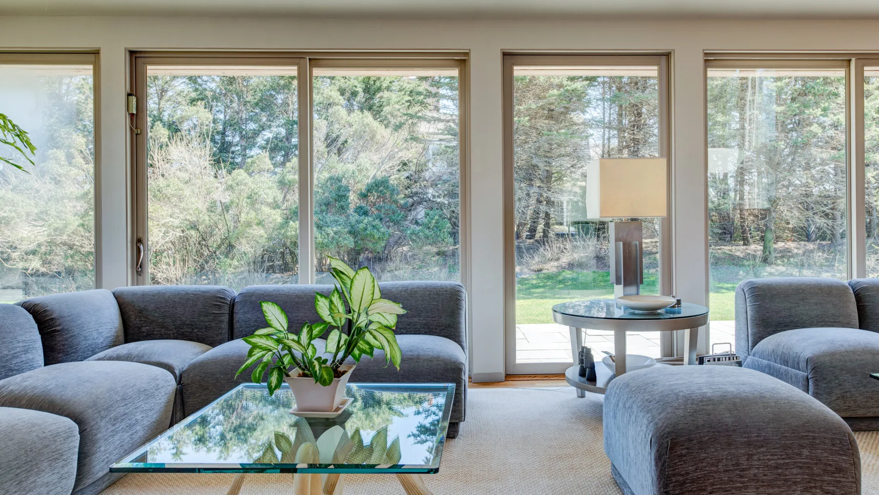 Una sala de estar con asientos grises y una mesa de centro de vidrio junto a grandes ventanas.