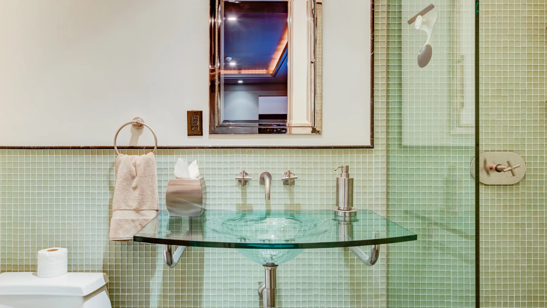 Un baño con una encimera de vidrio para el lavabo y paredes de azulejos verde pálido.