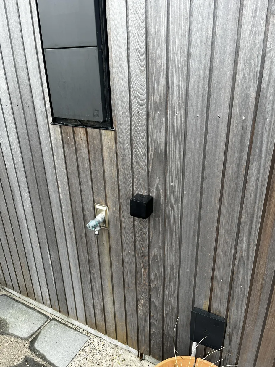 Una pared exterior revestida tiene una ventana pequeña, un grifo de agua y accesorios oscuros.