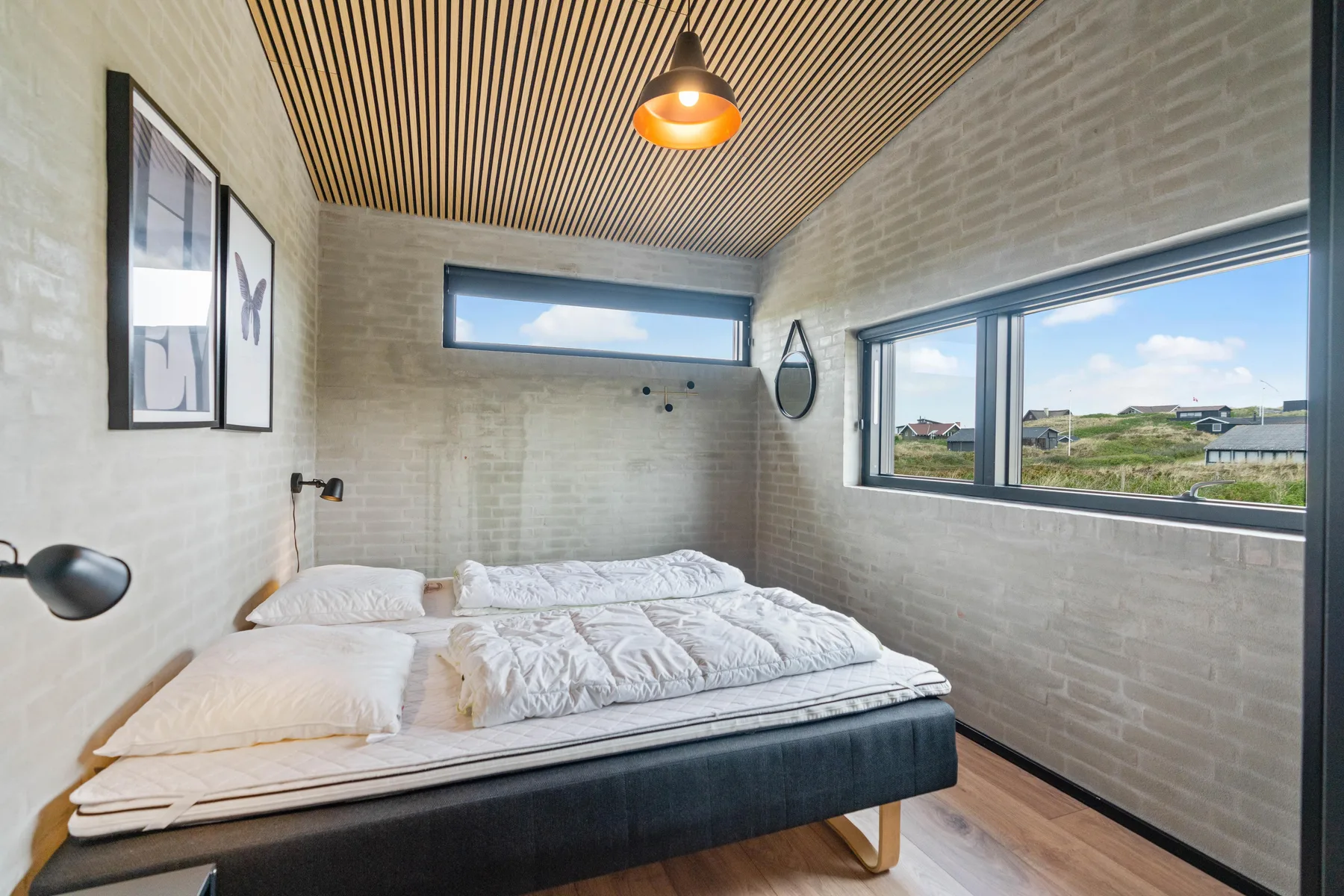 Un dormitorio con una cama bajo amplias ventanas con vistas a los edificios cercanos.