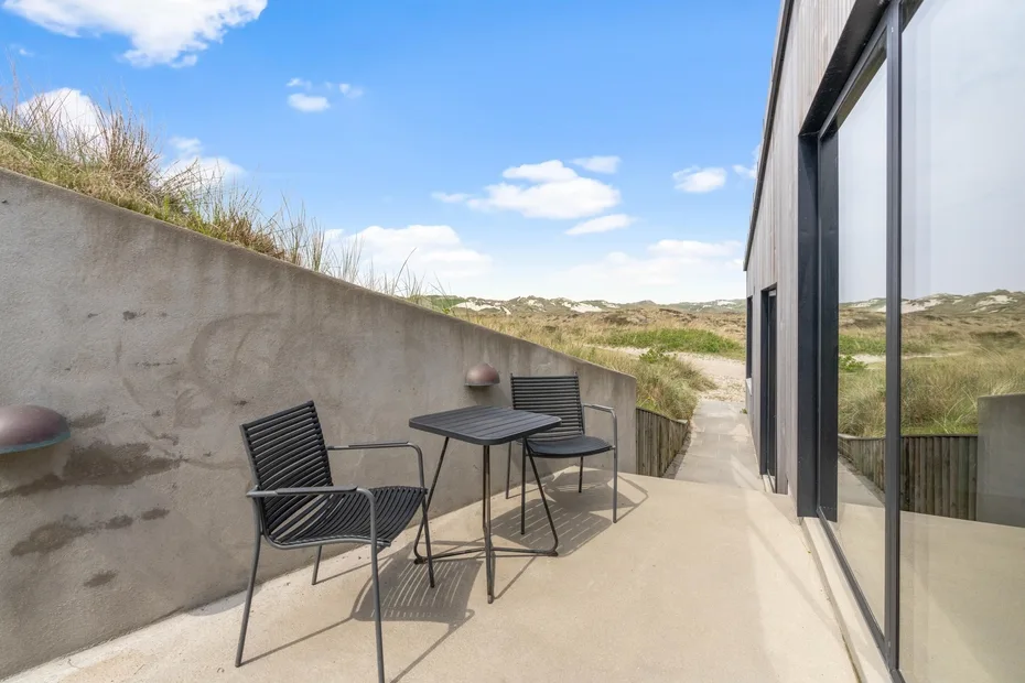 Un patio exterior con asientos junto a una pared de vidrio, frente a dunas cubiertas de hierba.